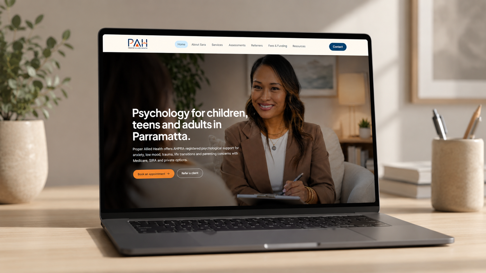 Laptop mockup displaying the Proper Allied Health website homepage on a desk in a softly lit office setting, with the hero section featuring a psychologist speaking with a client.